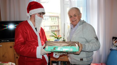 Na zdjęciu znajduje sie Mieszkaniec oraz Święty Mikołaj, który wręcza mu prezent. W tle wnętrze pokoju. Mieszkaniec uśmiecha się.