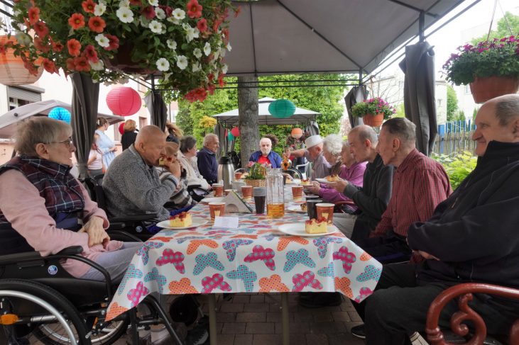 Na środku zdjęcia widać krótszy bok stołu, przy którym zasiada grupa starszych osób. Na stole znajdują się dzbanki z sokiem i talerze z ciastem truskawkowym. Ludzie przy stole uśmiechają się i rozmawiają. Stół przy którym siedzi grupa jest w ogrodzie, pod parasolem. Jest ładna pogoda.