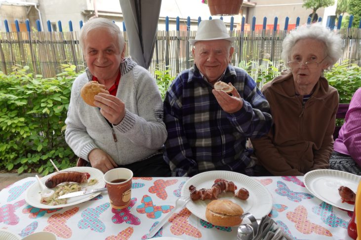 Na zdjęciu widać trzy starsze osoby siedzące przy ogrodowym stole. Przed nimi znajdują się talerze z upieczonymi kiełbaskami. Mężczyźni jedzą pieczywo, uśmiechają się, kobieta w okularach patrzy przed siebie.