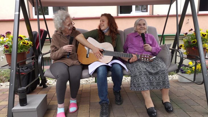 Na zdjęciu widać trzy osoby siedzące na ogrodowej huśtawce. Starsze kobiety po dwóch bokach śmieją się i śpiewają do mikrofonów. Młodsza kobieta w środku gra na gitarze i uśmiecha się do starszej kobiety po lewej stronie.