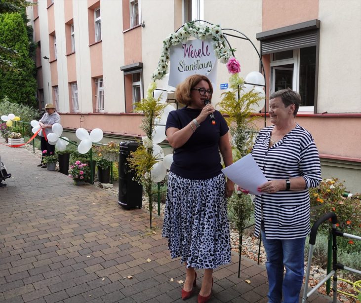 Dwie kobiety stoją w ogrodzie na tle budynku . Wzajemnie spoglądają na siebie i uśmiechają się. Jedna z kobiet trzyma w ręku mikrofon, do którego przemawia. Druga z kobiet w ręku trzyma kartkę papieru. Za nimi znajduje się udekorowana kwiatami i balonami pergola.