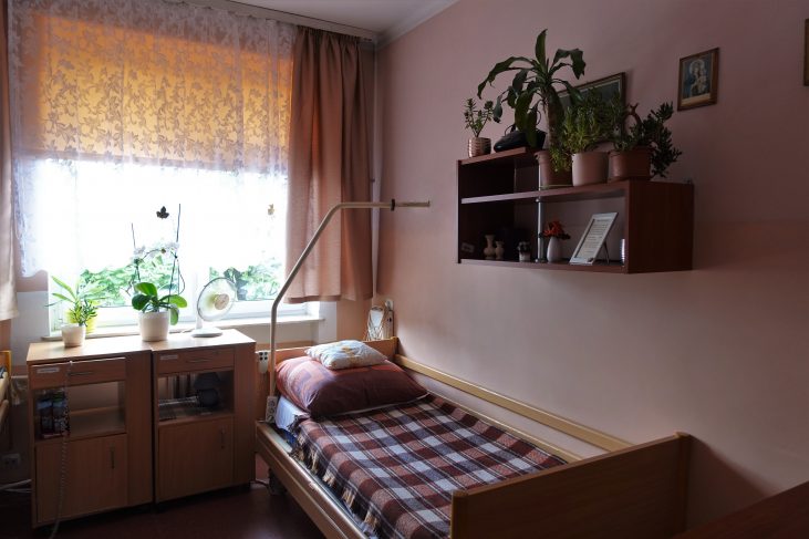 Pokój mieszkalny w Domu Pomocy Społecznej. Przy ścianie stoi łóżko z uchwytem dla osób niepełnosprawnych, nad łóżkiem znajduje się wisząca szafka. Po lewej stronie znajdują się szafki przyłóżkowe.