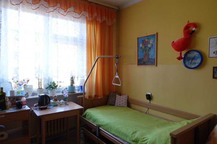 Pokój mieszkalny. Przy ścianie znajduje się łóżko zaścielone zieloną narzutą. Do łóżka jest przymocowany uchwyt. W oknach wiszą pomarańczowe zasłony. Po lewej stronie znajduje się stolik. Nad łóżkiem są zawieszone: obraz, pluszowy flaming i zegar