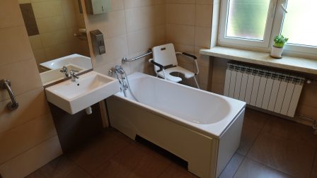 Łazienka w Domu Pomocy Społecznej. Znajduje się w niej wanna, umywalka, lustro, krzesło kąpielowe, uchwyty dla osób niepełnosprawnych przytwierdzone do ścian.