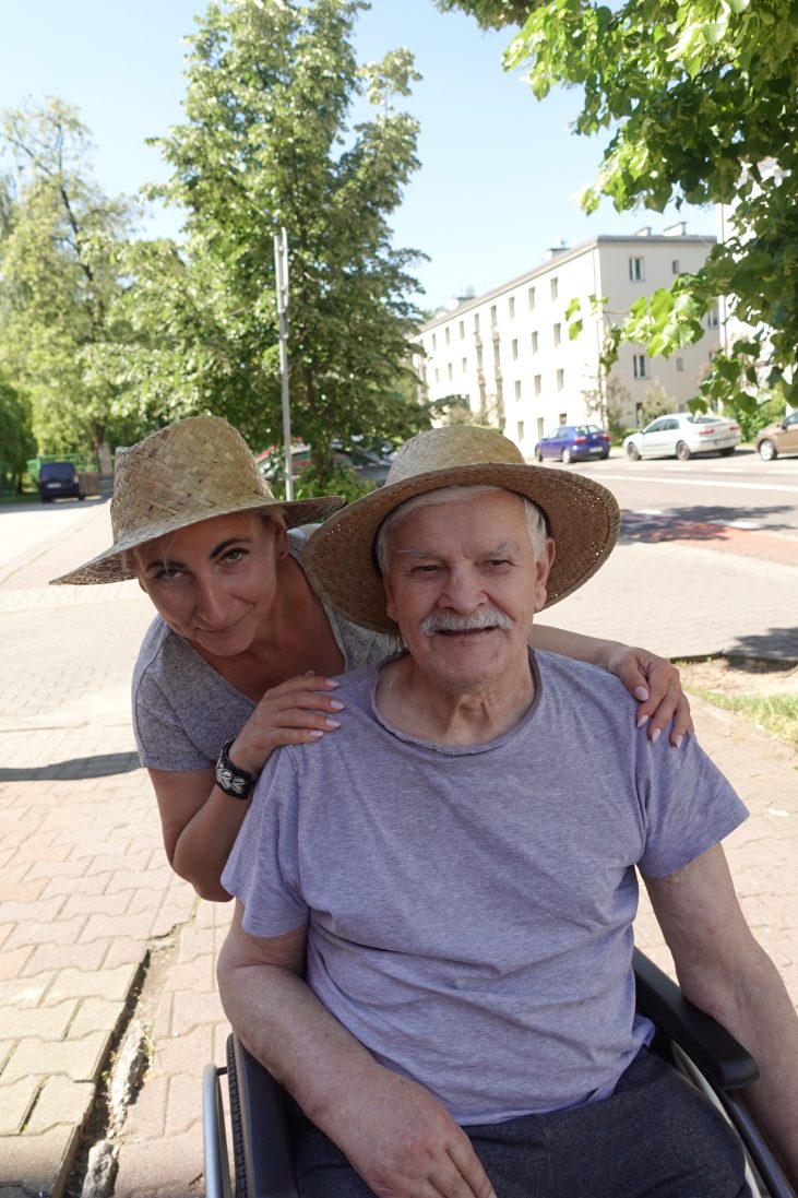 Kobieta w średnim wieku i Starszy mężczyzna w słomianych kapeluszach uśmiechają się. Kobieta trzyma mężczyznę za ramiona, w tle widać ulicę i budynki.