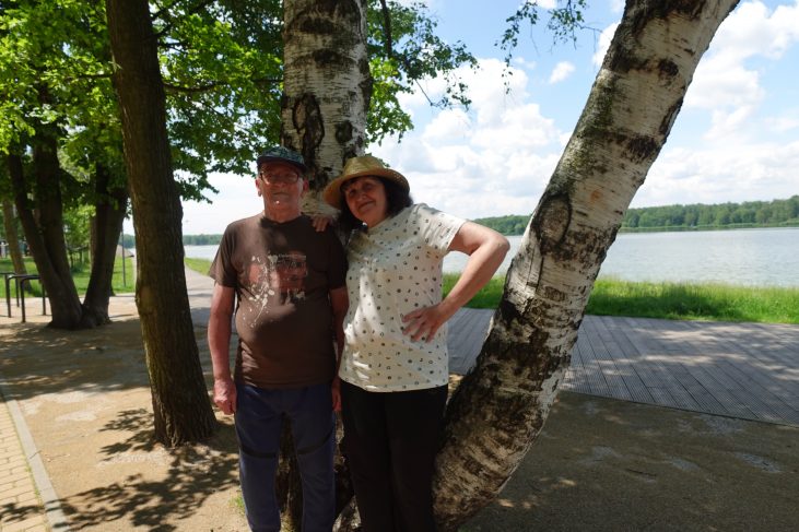 Starszy mężczyzna i kobieta pozują do zdjęcia. Kobieta uśmiecha się , rękę ma położoną na ramieniu mężczyzny. W tle jest są drzewa, trawa, jezioro.