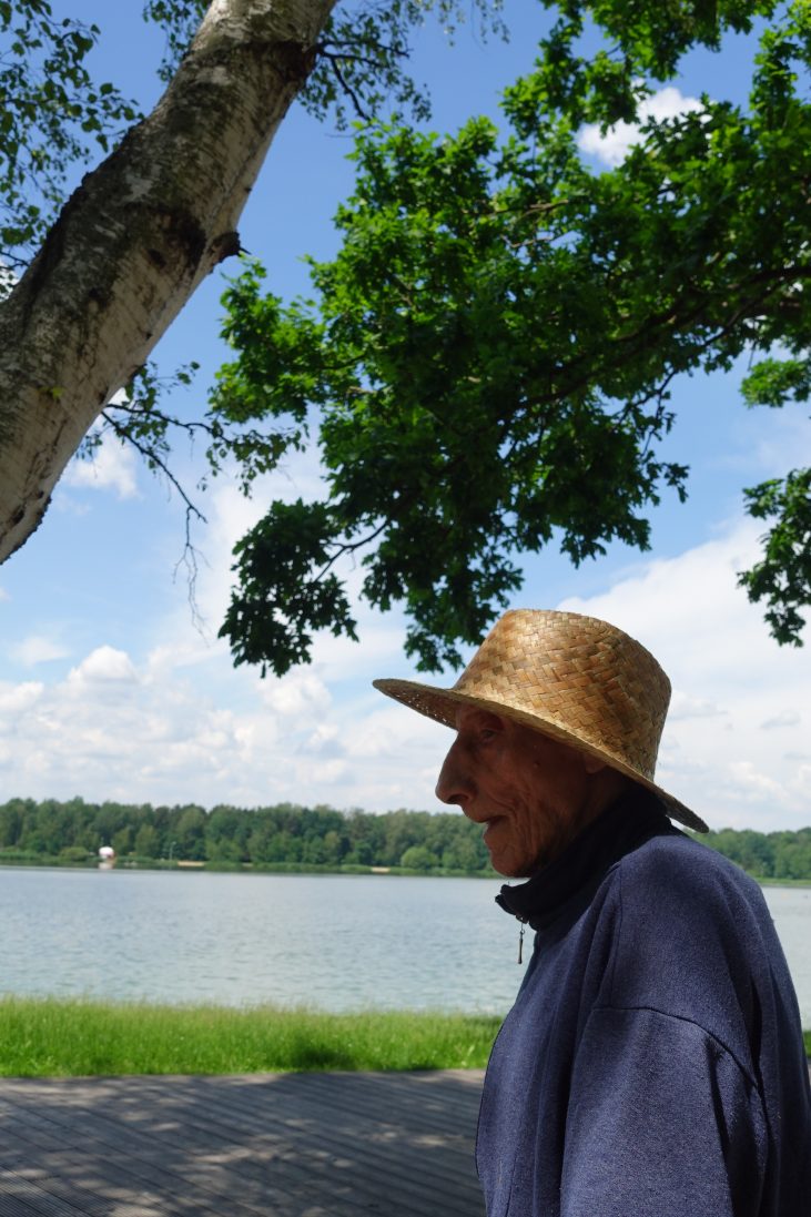 Na pierwszym planie widać profil twarzy starszego mężczyzny w słomianym kapeluszu. Nad nim jest drzewo, a w tle znajduje się duże jezioro i niebieskie niebo