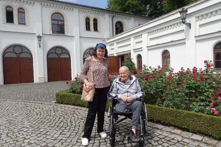 Starsza kobieta uśmiecha się, rękę ma położoną na ramieniu starszego mężczyzny który siedzi na wózku. W tle jest uprawa różowych róż oraz biały budynek.