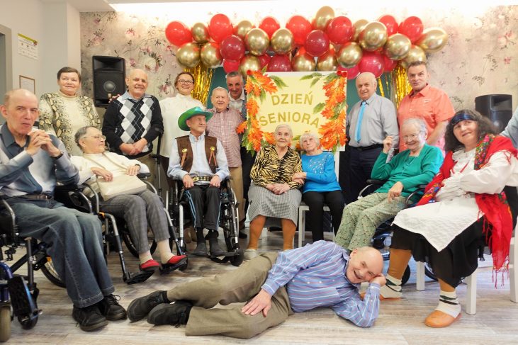 Piętnaście osób w różnym wieku, niektórzy z nich na wózkach inwalidzkich znajdują się w jadalni Domu Pomocy. Na pierwszym planie leży mężczyzna i szeroko się uśmiecha. Wszyscy cieszą się. W tle widać złota kurtynę i bordowo-złote balony. W centrum znajduje się plakat na którym jest napisane Dzień Seniora, udekorowany jesiennymi liśćmi.