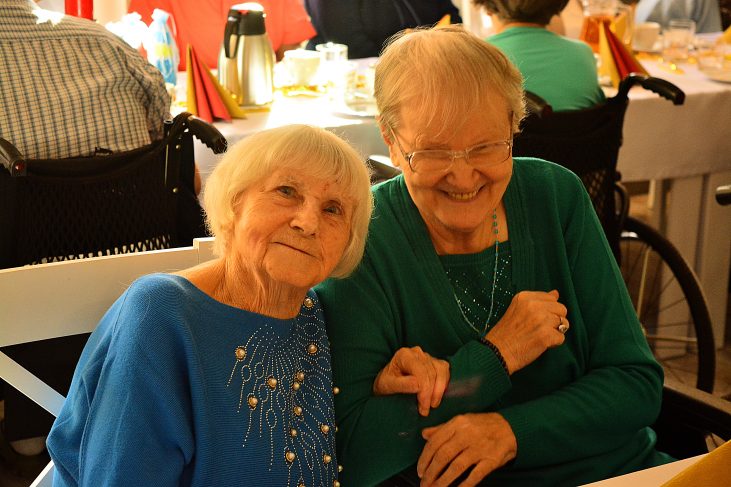 Dwie starsze kobiety siedzą na biesiadzie przy stole. Obejmują się nawzajem i uśmiechają.