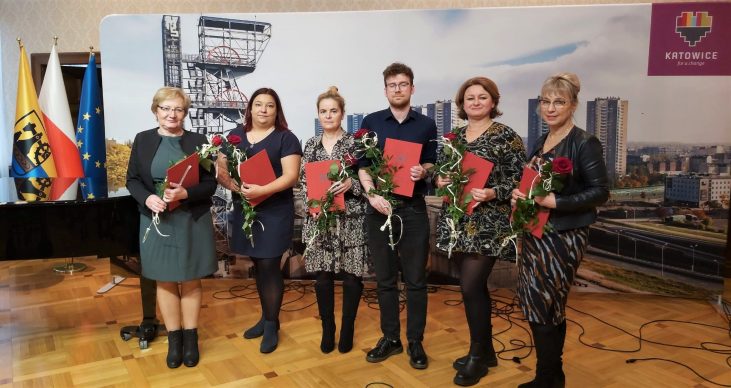 2. Pięć kobiet i jeden mężczyzną stoją w rzędzie, trzymają czerwone okładki i róże. Po lewej stronie znajduje się flaga Śląska, Polski oraz Unii Europejskiej. W tle panorama Katowic