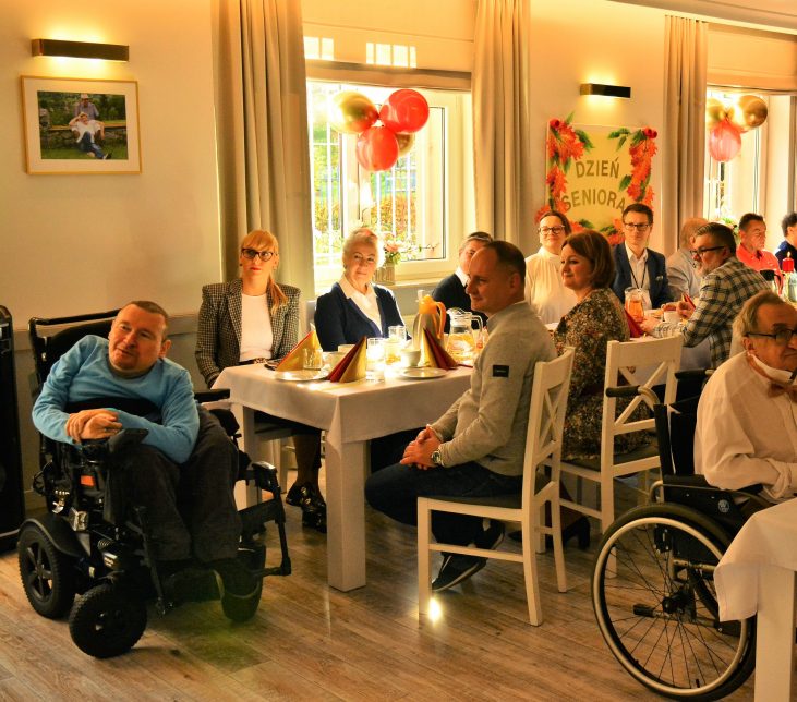 Grupa gości siedzi przy odświętnie nakrytym stole podczas uroczystości z okazji Dnia Seniora. stół znajduje się przy oknie. W pierwszym planie widać mężczyznę, który siedzi na wózku elektrycznym dla osób niepełnosprawnych. Mężczyzna uśmiecha się. Inne osoby patrzą przed siebie, oglądają występy i uśmiechają się. W tle na ścianie wisi duży napis "Dzień Seniora"