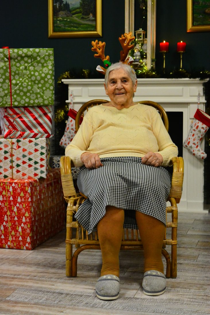 Starsza kobieta z opaską z uszami renifera siedzi na drewnianym fotel u i uśmiecha się. Po lewej stronie znajduje się góra prezentów. W tle widać kominek, na którym są czerwone świece na złotych świecznikach oraz stroik