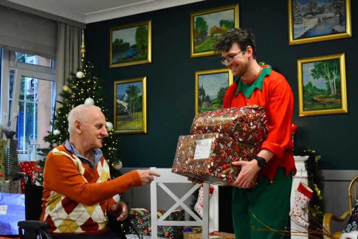 Starszy mężczyzna w pomarańczowym swetrze uśmiecha się i wyciąga rękę. Młody mężczyzna w stroju elfa szeroko się uśmiecha i daje czerwony prezent. W tle choinka, prezenty, biały kominek.