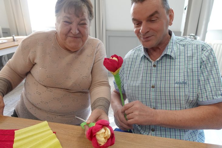 W jadalni DPS siedzi z lewej strony kobieta a z prawej mężczyzna. Mężczyzna i kobieta trzymają po jednym kwiatku z krepiny uśmiechają się. Mężczyzna ma spuszczony wzrok w dół a twarz skierowaną w lewo, a kobieta patrzy na wprost.