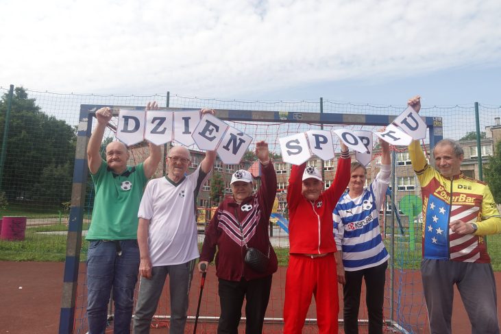 Na boisku przed bramką piłkarską stoi 6 osób, 2 kobiety i 4 mężczyzn. Trzymają napis "dzień sportu" unosząc ręce. Uśmiechają się.