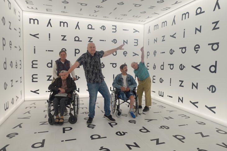 W Muzeum Śląskim pokoju, w którym dookoła znajdowały się litery znajdują się 2 kobiety i 3 mężczyzn. Uśmiechają się. Mężczyzna po środku oraz z prawej strony zdjęcia pochylają się z rozłożonymi rękami na kształt jaskółki.