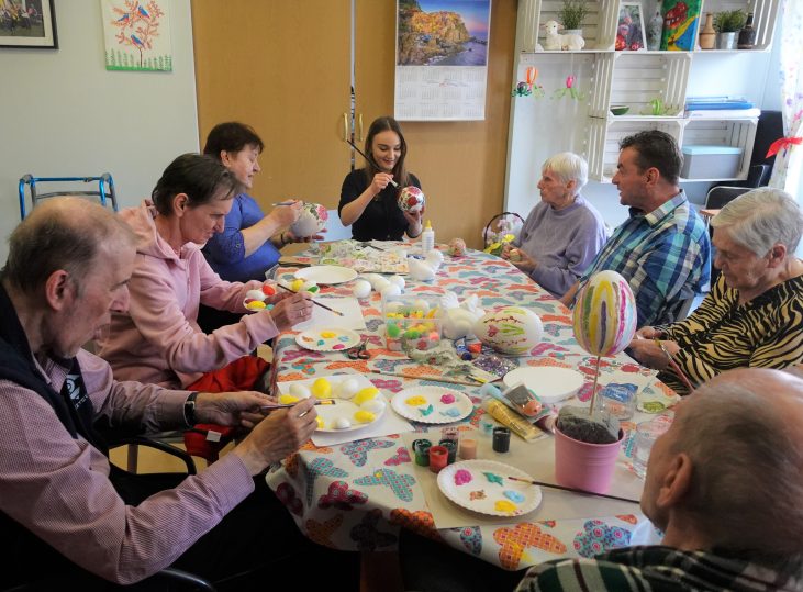 Grupa starszych osób siedzi przy stole. Kobiety i mężczyźni malują jajka wielkanocne.