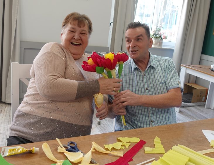 Dwóch seniorów siedzi przy stole. Pan trzyma piękne kwiaty zrobione z papieru i uroczo podarowuje je kobiecie obok. Kobieta przyjmuje bukiet i promiennie się uśmiecha.