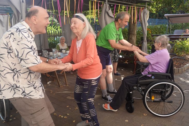 Seniorzy trzymają się za ręce i tańczą z radością. W tle możemy zobaczyć starszego mężczyznę tańczącego z uśmiechniętą kobietą na wózku.